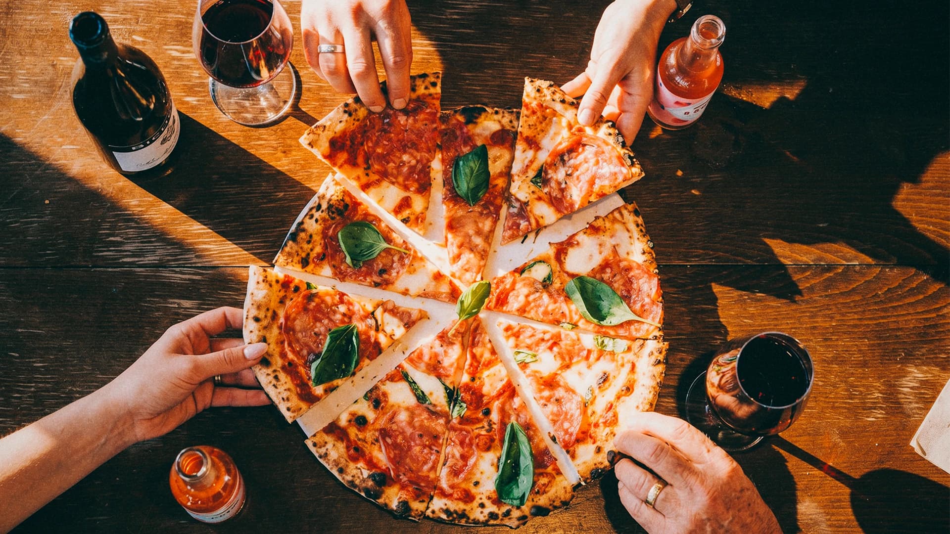 Ylhäältä kuvattu pizza jaettuna neljään osaan ja neljä kättä ottamassa siivua